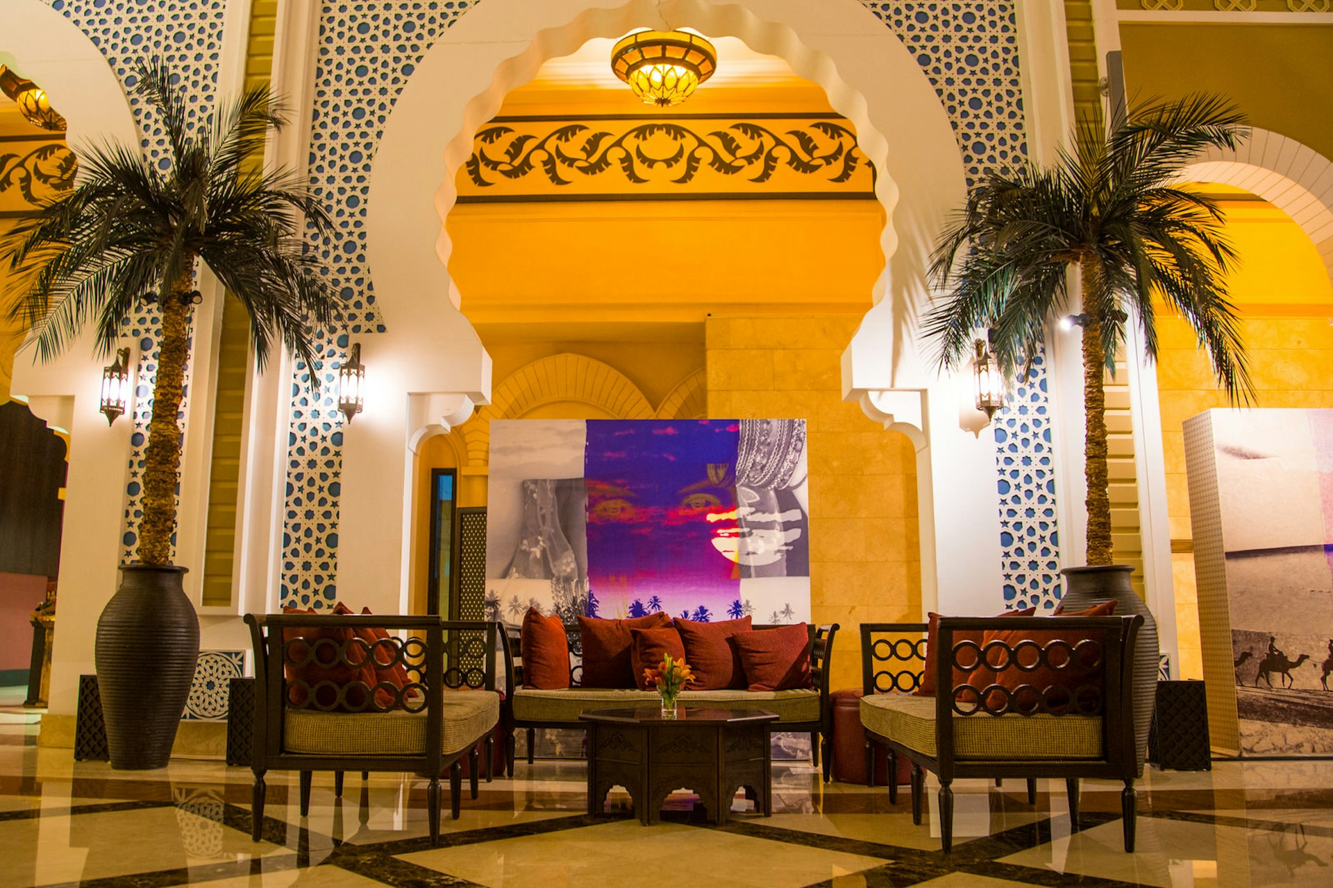 Hotelllobby i Dubai, Förenade Arabemiraten.  Träsoffor med djupröda kuddar är arrangerade under en utarbetad valvgång genom vilken vi kan se lite modern konst.  Valvet flankeras av höga, krukväxta palmer. 