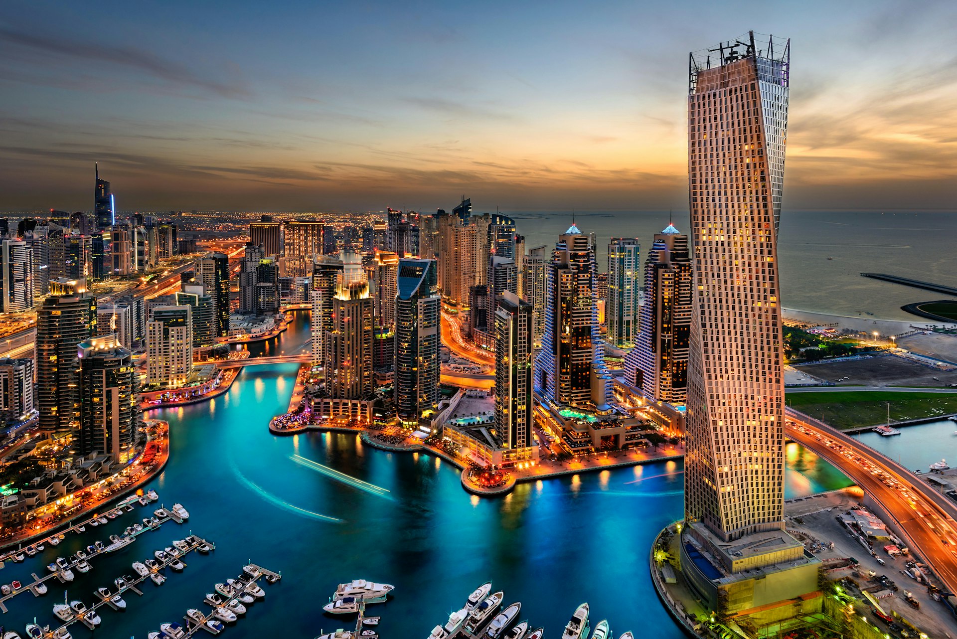 En långexponerad bild av Dubai Marina, Förenade Arabemiraten i skymningen, tagen från högt upp i en av de många skyskraporna.  Det ligger massor av båtar som ligger till kaj i marinan, gatorna är upplysta liksom de olika skyskraporna. 