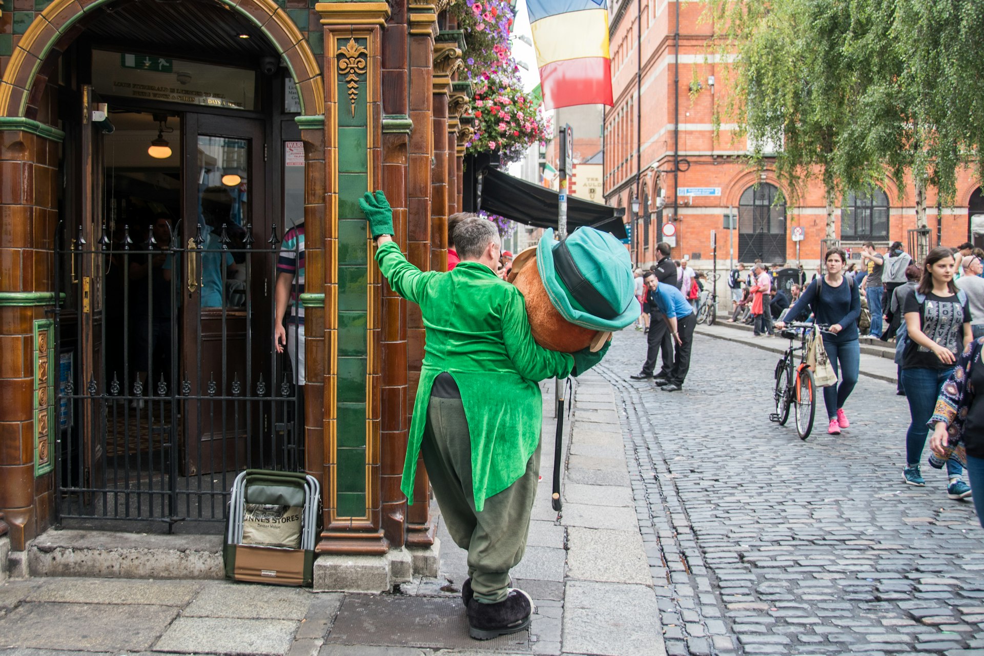 A man in a huge leprechaun outfit with his back to the camera, leaning against a tiled wall. The man has taken the giant head off the costume while he rests on the cobbled street