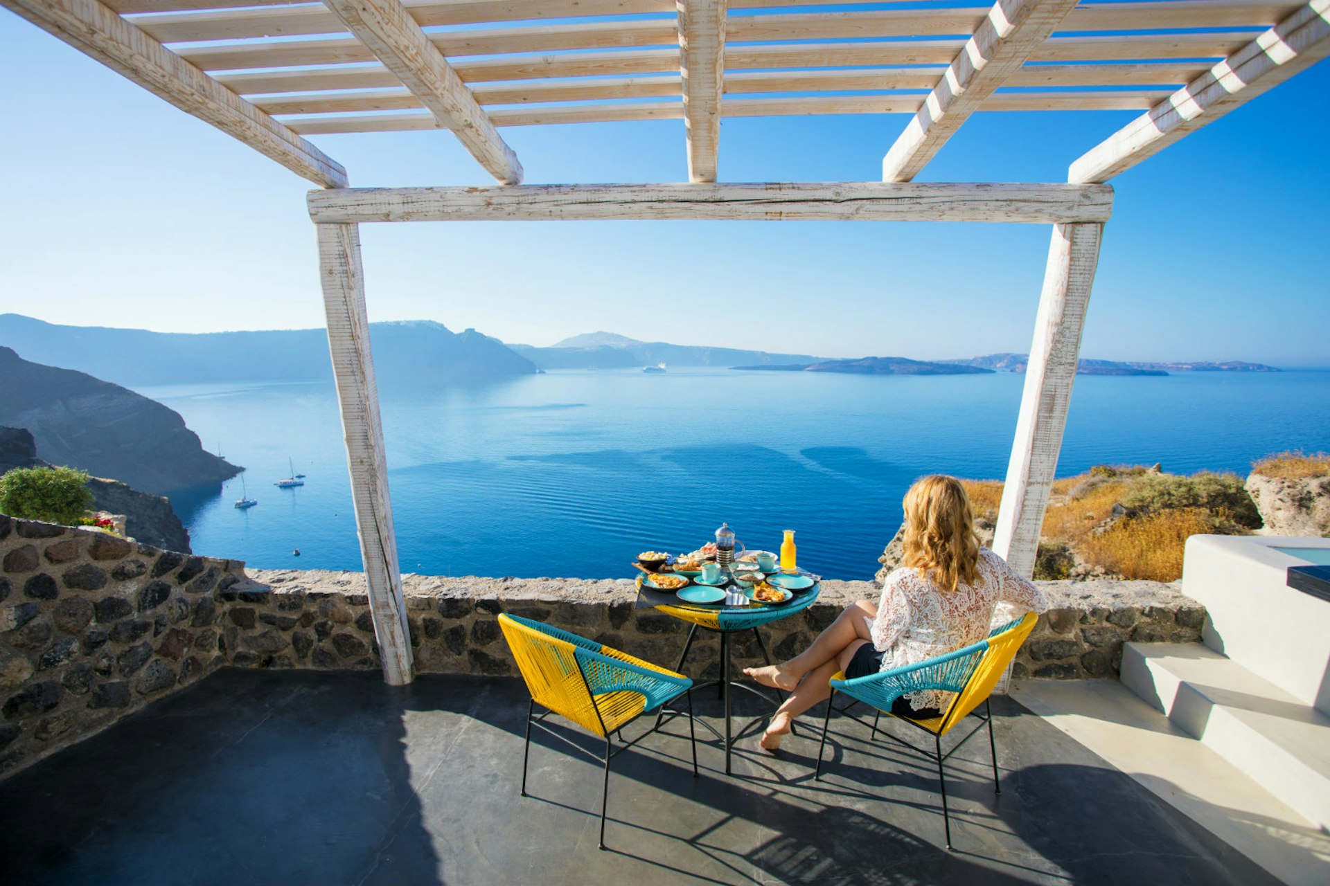Romantic trip with kids in tow – A woman looks out over the Santorini coastline from the breakfast table.