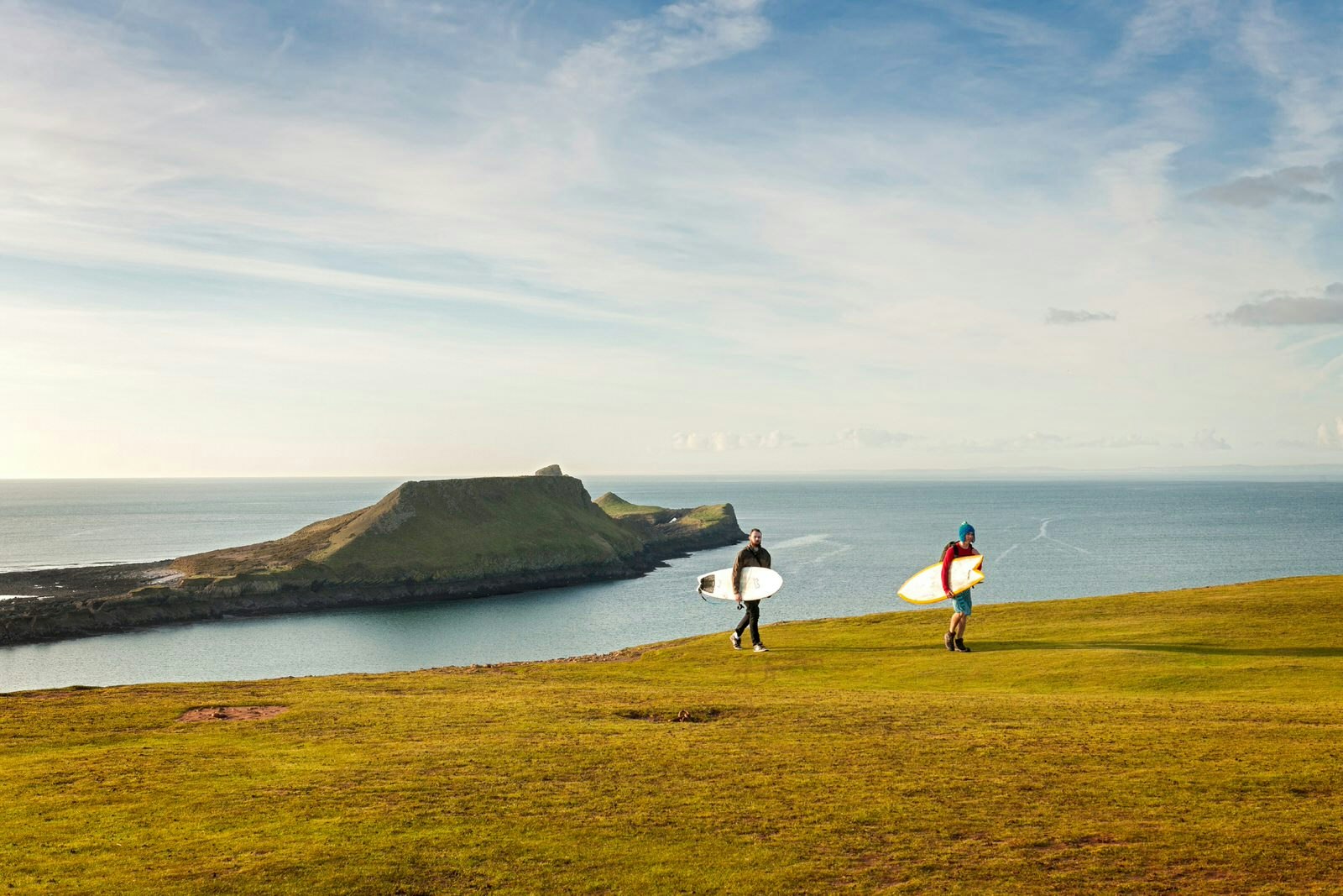 Surfers return from the water on Wales's Gower Peninsula, with Worm's Head in the background © VisitWales