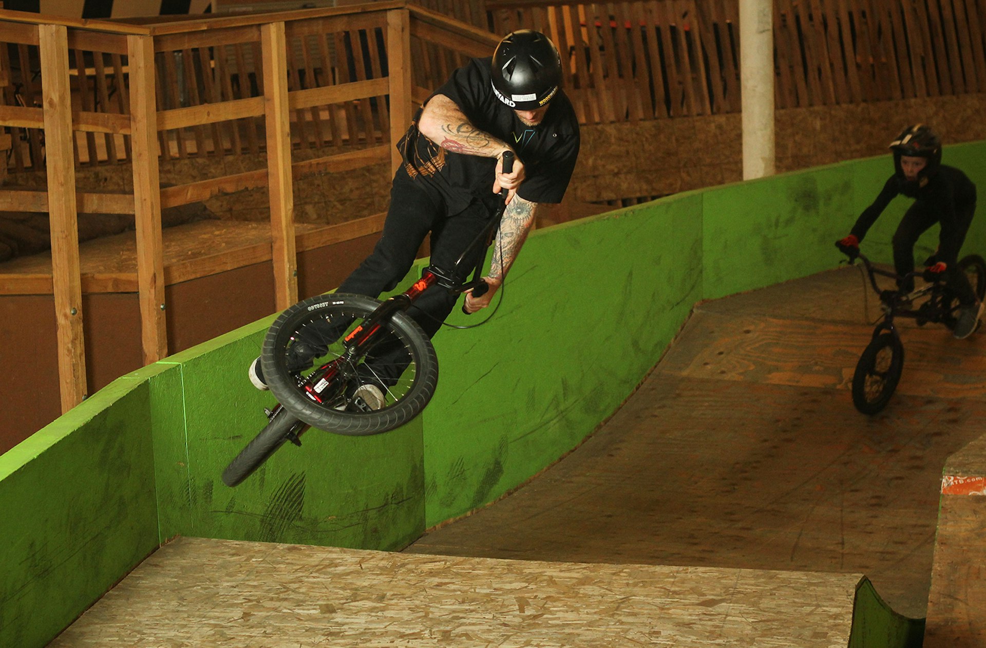 A man dressed in all black with tattooed arms jumps over a wooden ramp on a BMX bicycle at an indoor track. Another rider is coming up behind him to make the same jump.