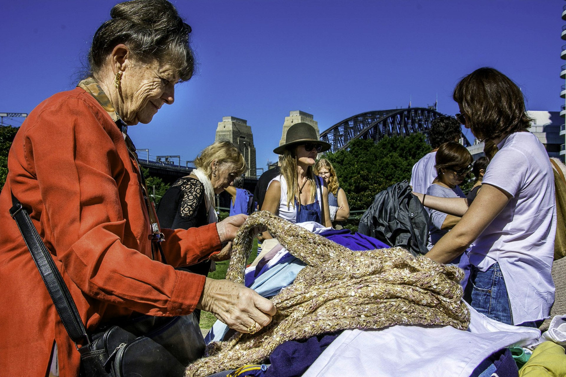 A lady examines a woolly jumper at Kirribilli Market in front of Sydney Harbour Bridge and a lovely blue sky