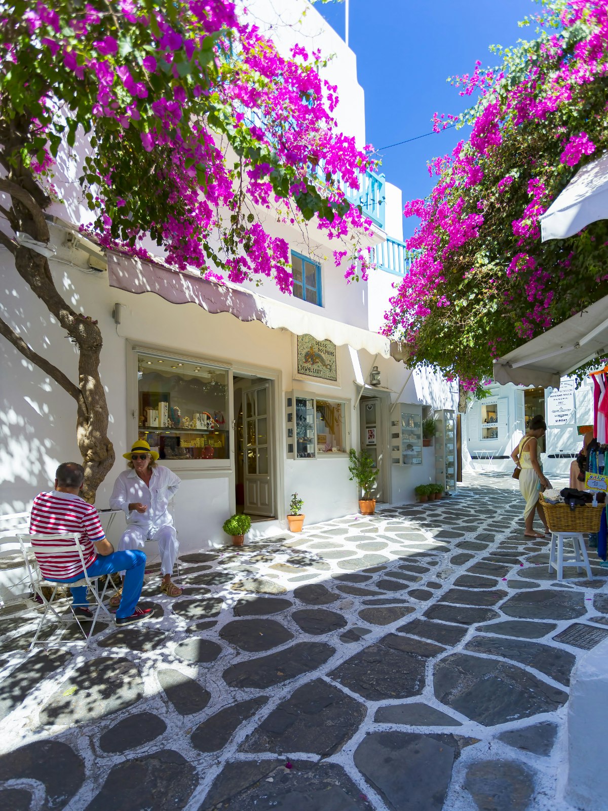 Small street lined with purple bougainvillea, shop fronts and small cafes. There are seats outside some of the cafes with people sitting and talking in Hora, Mykonos, Greece