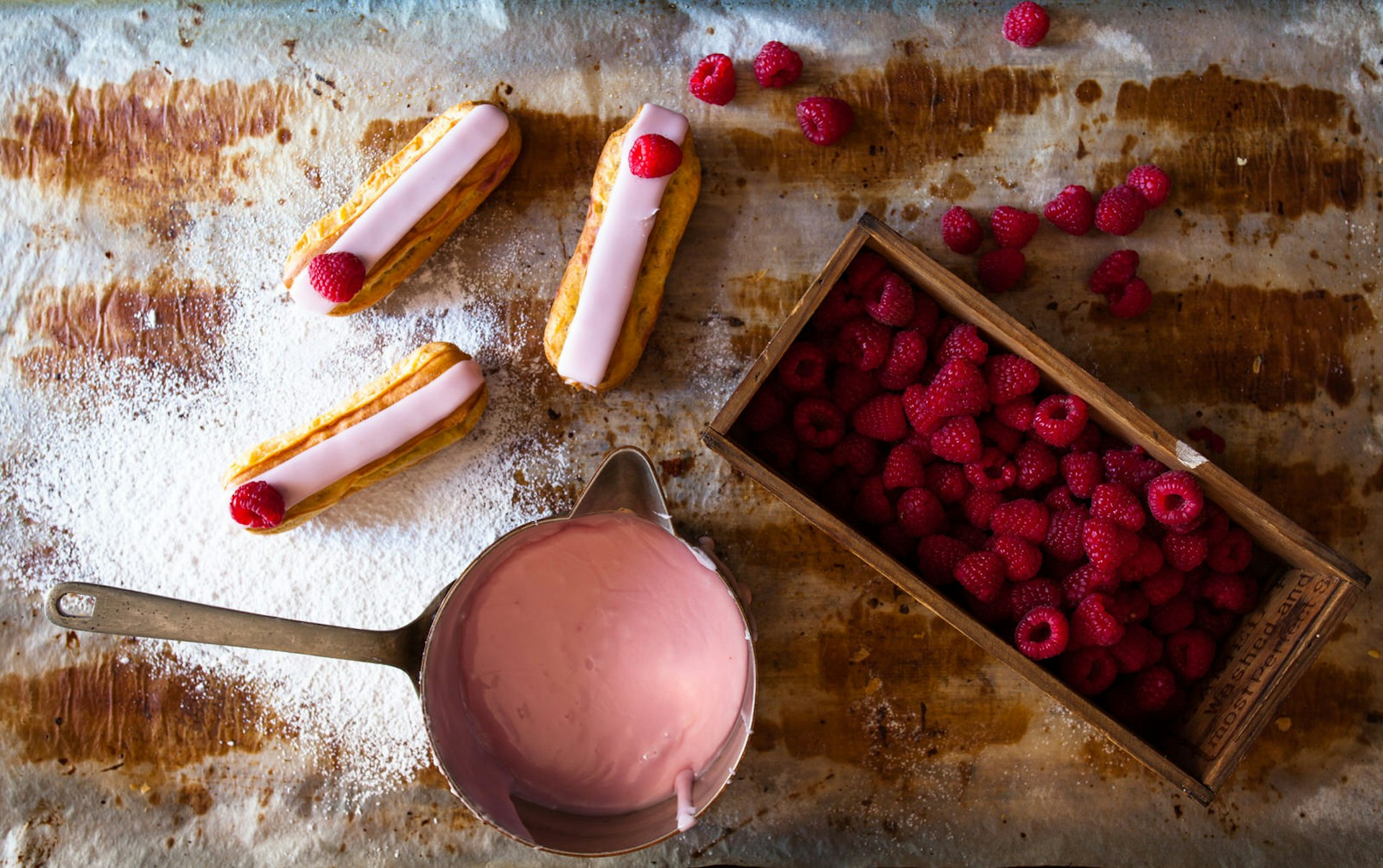 Raspberry eclairs sit next to some pink icing and fresh raspberries on a sugary table
