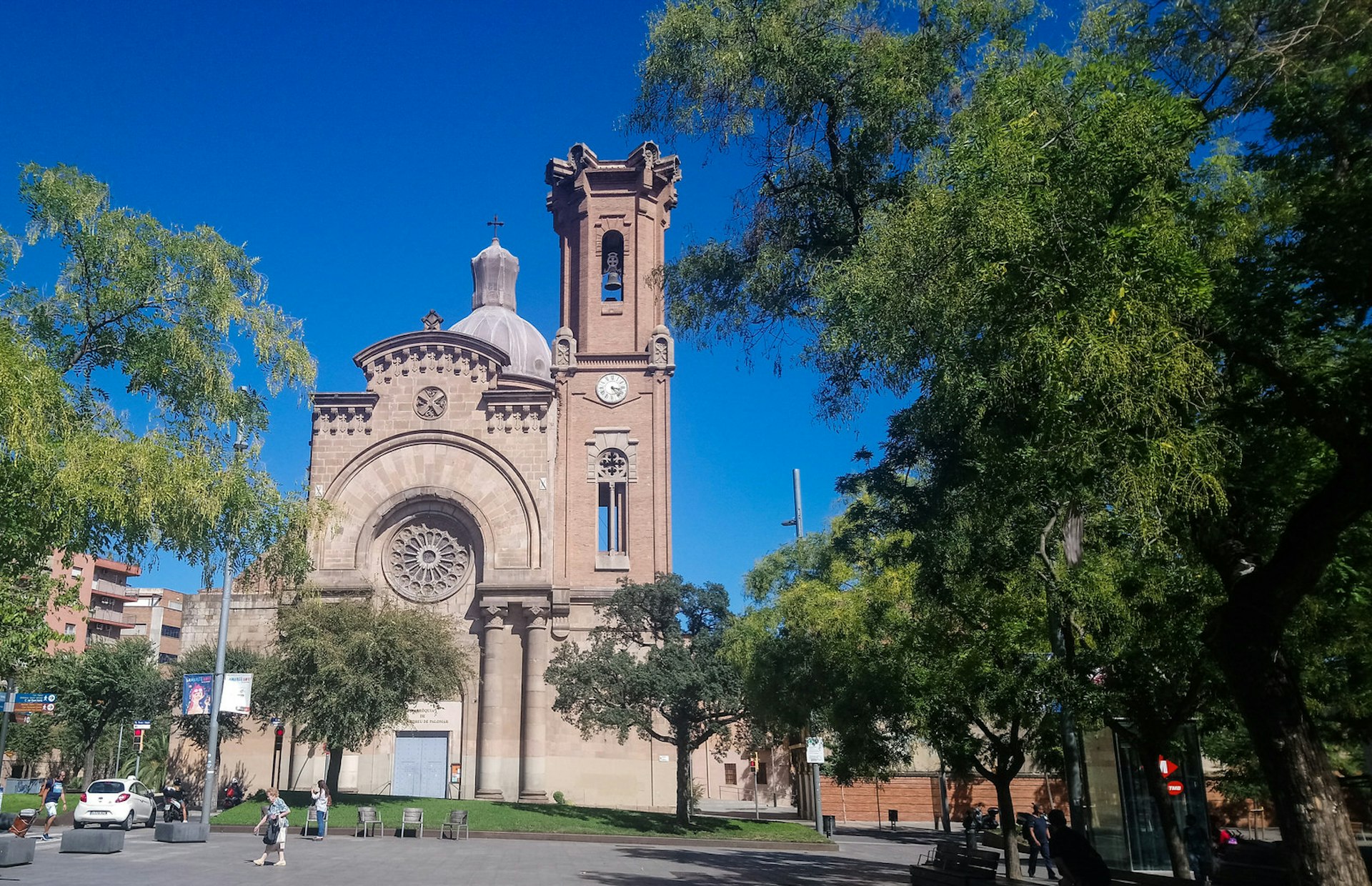 The pink-hued, neo-Gothic Church of Sant Andreu in Barcelona; the church has a tower and a rose window and sits in a leafy square.