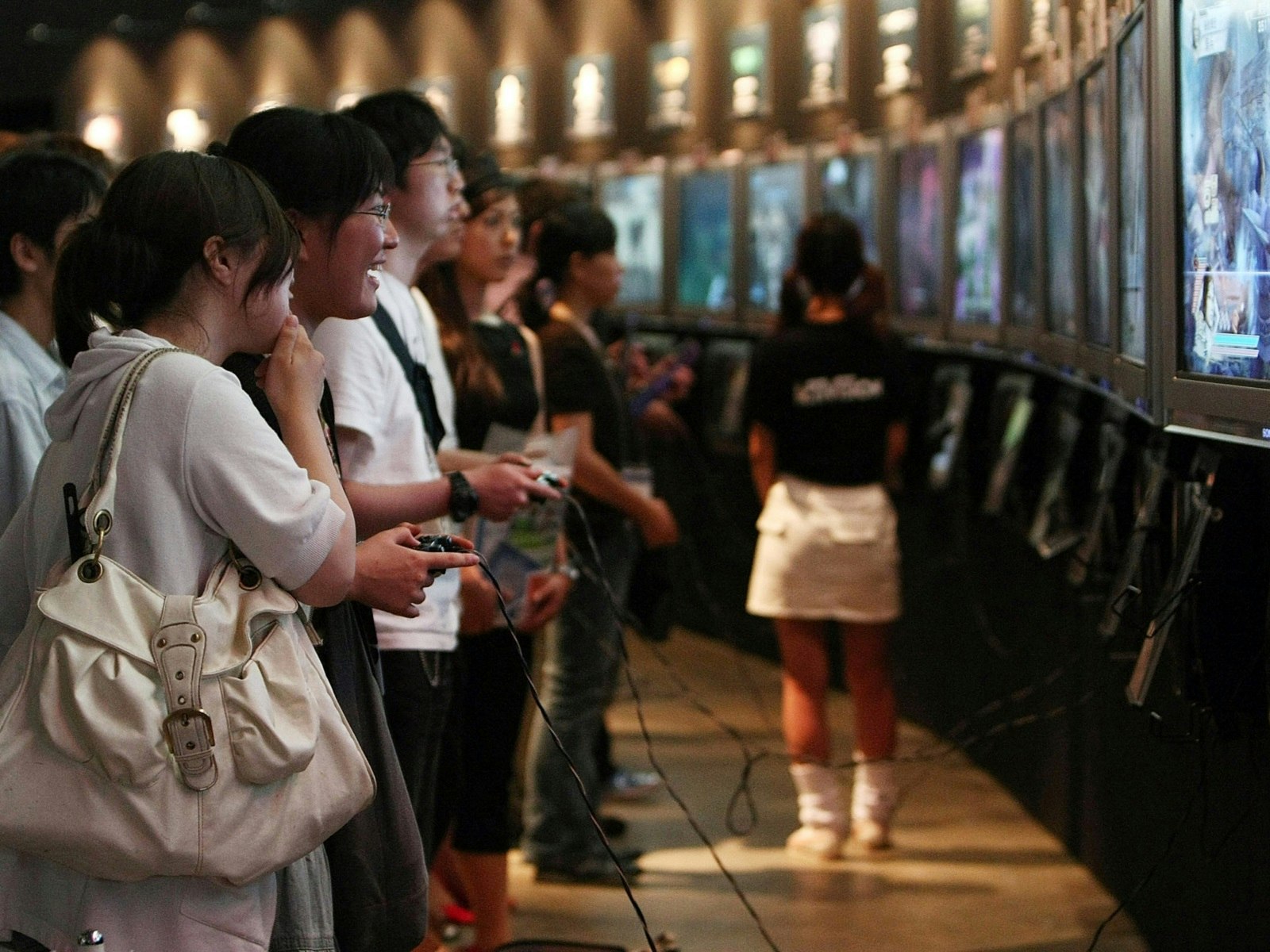 A group of people hold game controllers while standing in front large screens; Tokyo