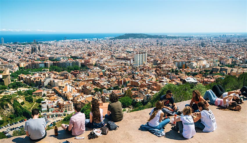 A group of young people sit on top of one of the concrete Bunkers del Carmel; below them unfolds a view over Barcelona, with the bright blue Mediterranean in the distance.