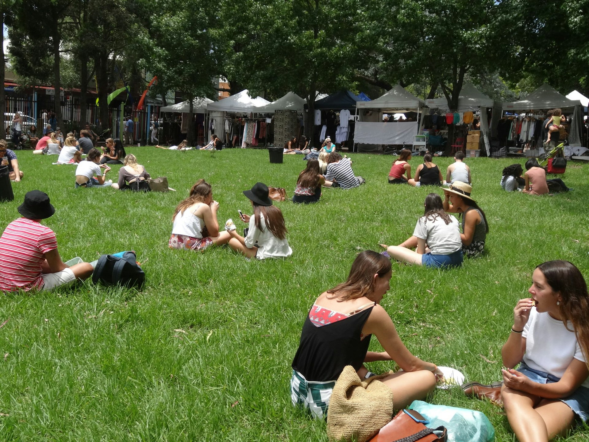 People sit on a wide open green space eating the food they've bought from market stalls in the the background