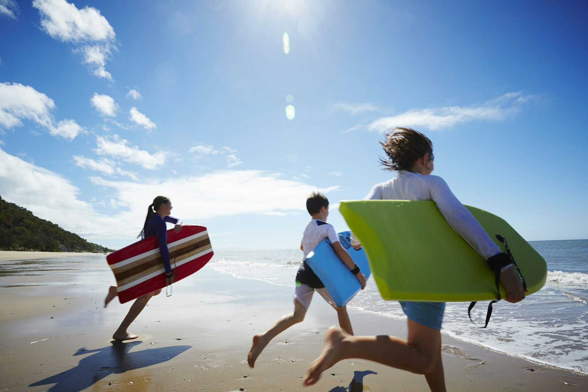 Three kids dressed in swimsuits and rash vests run into the sea holding colourful bodyboards 