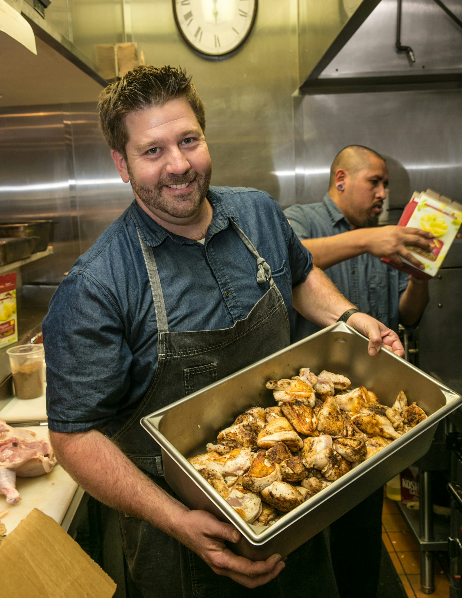 Dustin Valette smiles as he shows the camera a tray of chicken thighs while behind him, the line cook continues to prepare food for firefighters and first responders