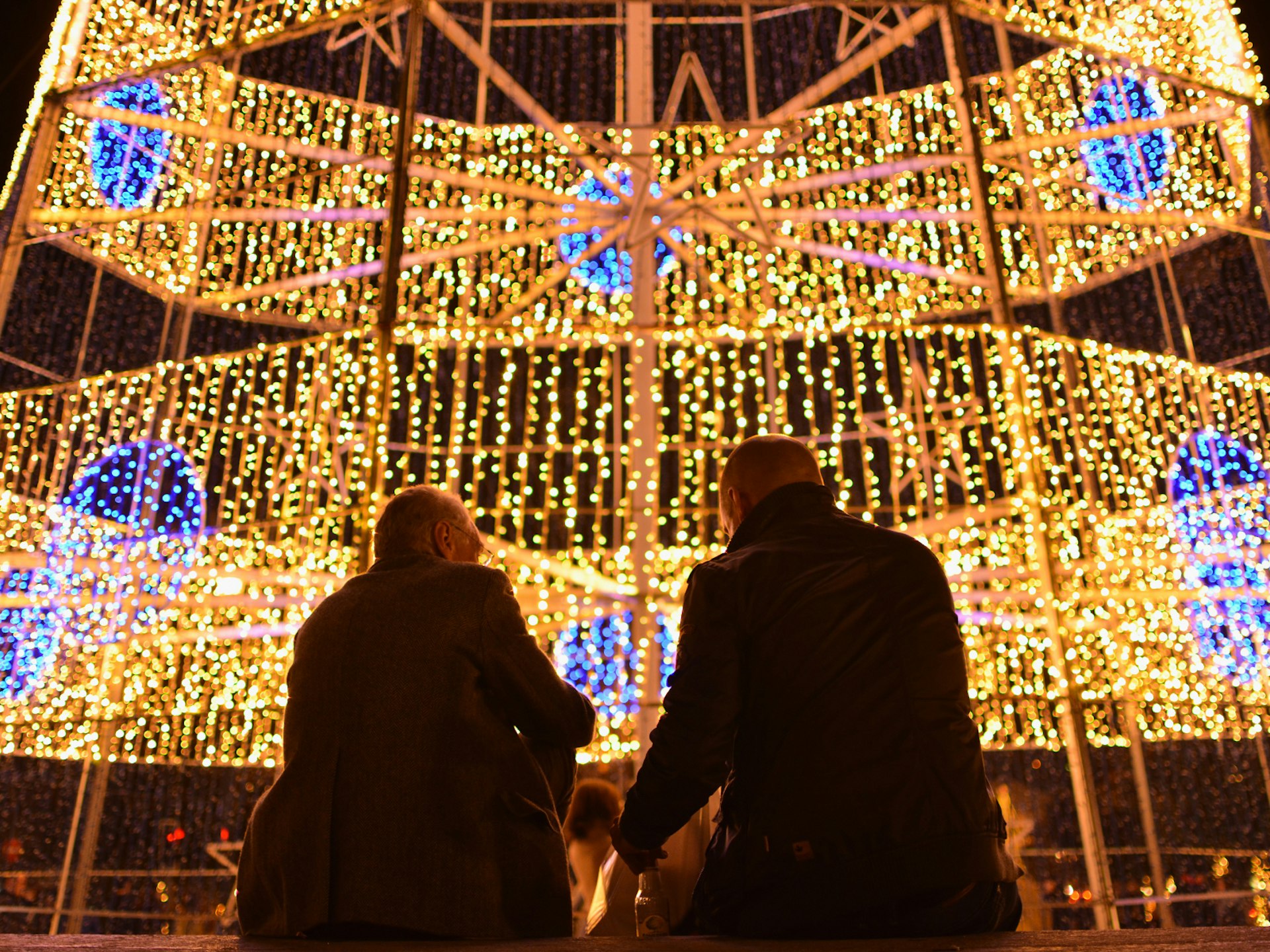 Two men sit in front of a Christmas tree made of lights in Porto, Portugal
