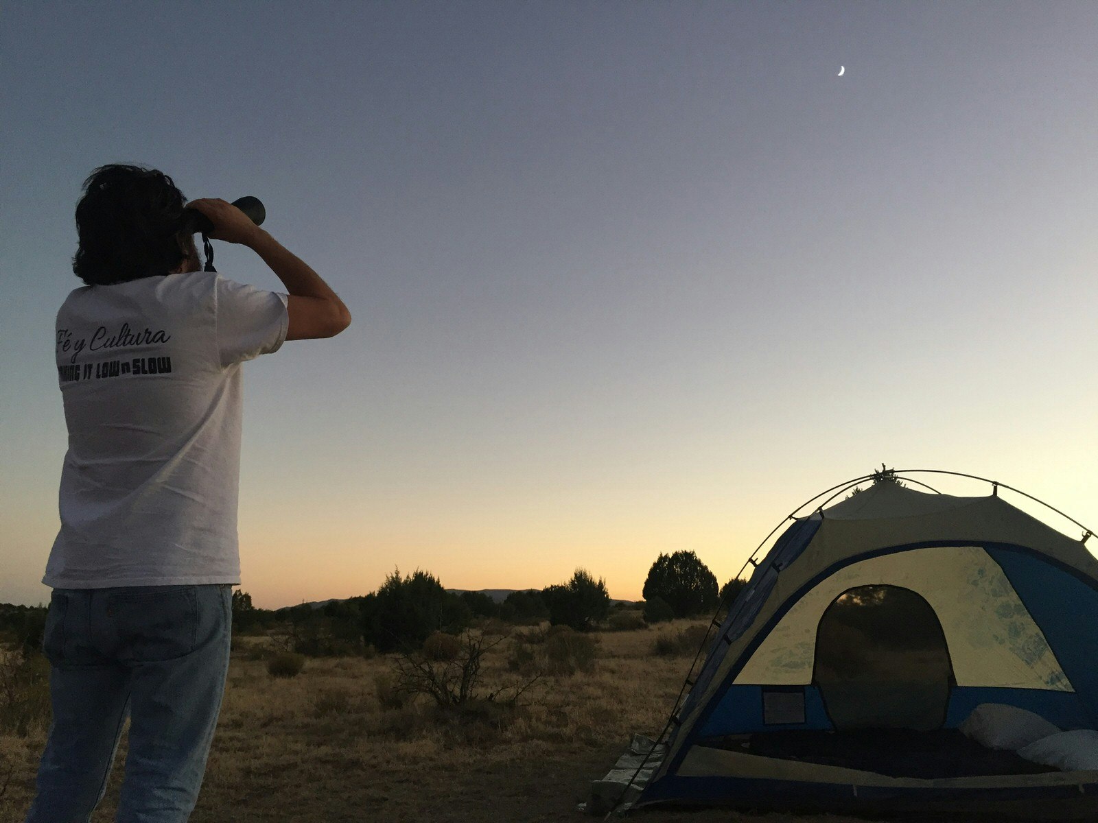 A man in a white t-shirt holds a pair of binoculars looking at a crescent moon at dusk next to a tent in the desert; Stargazing Southwest USA