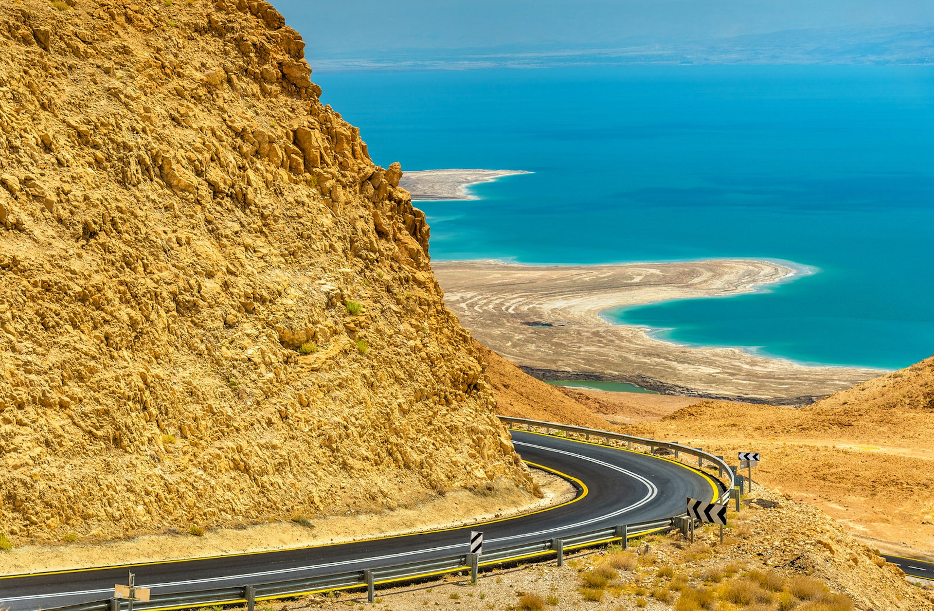 Road in the Judean desert rounds a bend that hugs an orange cliff and leads to the Dead Sea in the distance below..