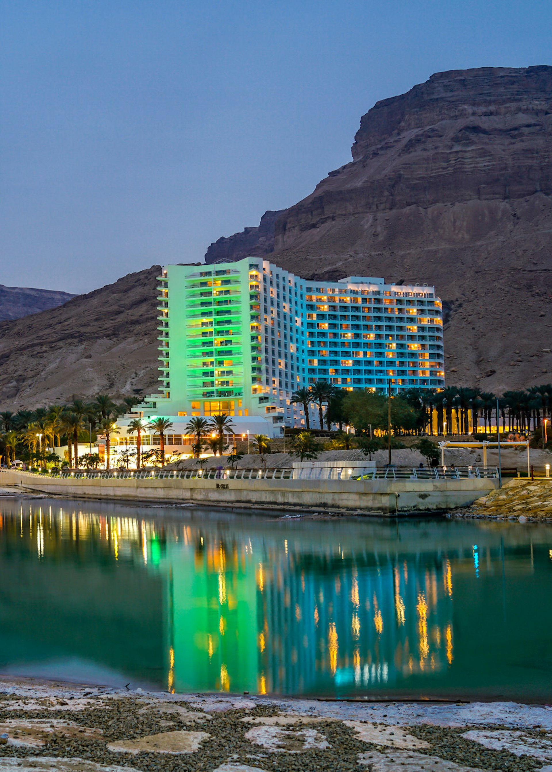 A white hotel resort glows green in mood lighting; it's backed by steep rocky cliffs and is fronted by the waters of the Dead Sea.