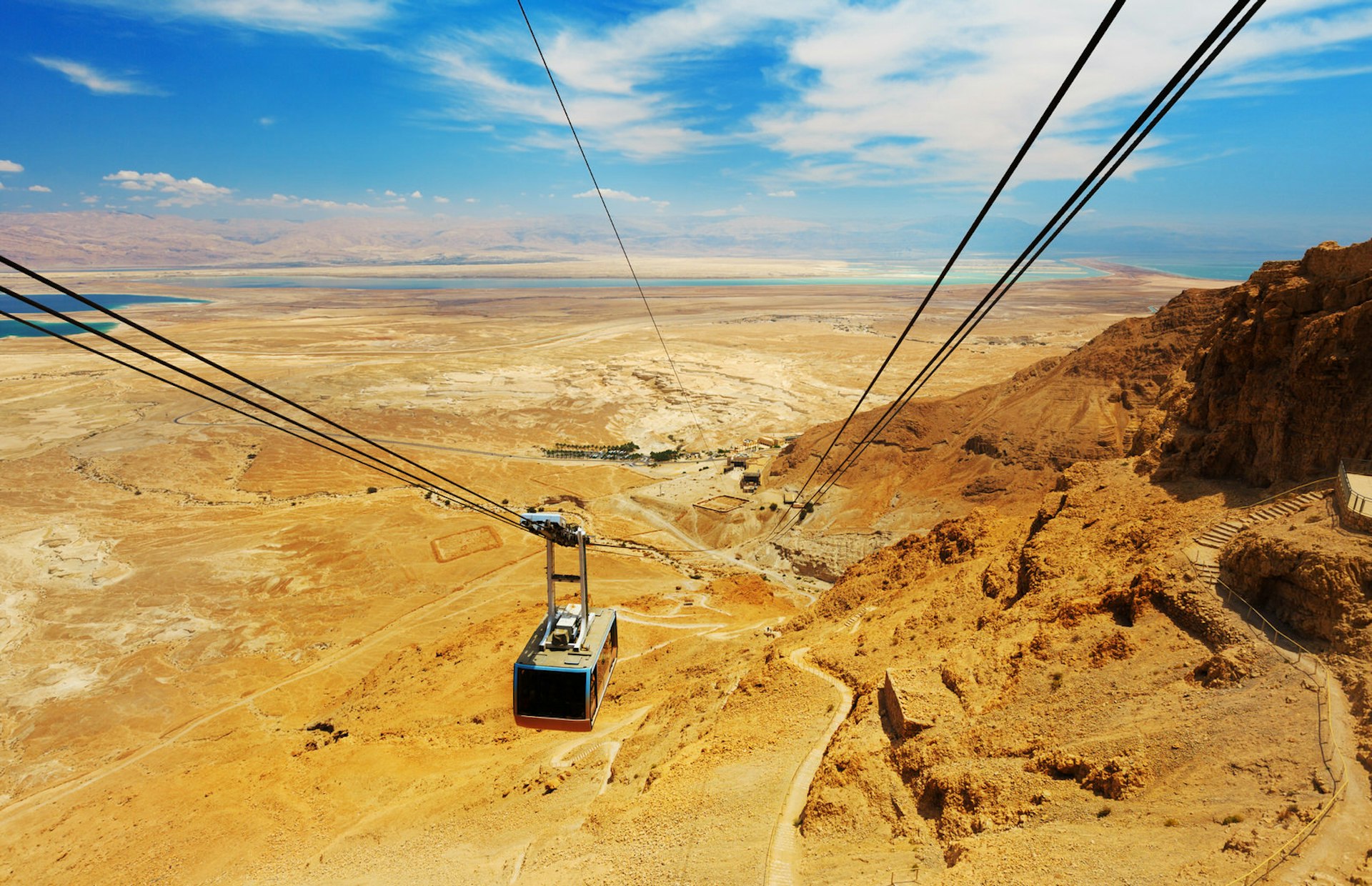 Masada cable car descends from the sky down to an orange landscape around the distant lake.