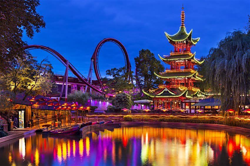 Le parc d'attractions des jardins de Tivoli, Copenhague;  on voit un lac au premier plan reflétant les illuminations d'une pagode de style japonais et des montagnes russes avec des boucles.