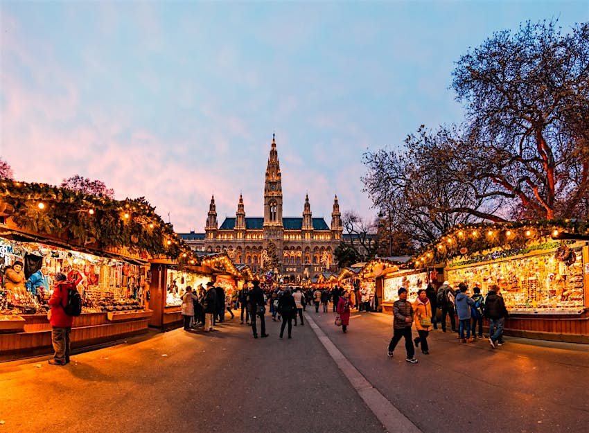 Marché de Noël au coucher du soleil à Vienne, Autriche;  les acheteurs parcourent les étals de chaque côté de la route, tandis qu'à la fin se trouve un bâtiment imposant avec des flèches.