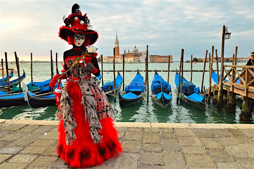Fêtard masqué vêtu de rouge, blanc et noir à Venise, en Italie, avec une rangée de gondoles derrière eux et les toits de la ville de l'autre côté de l'eau.