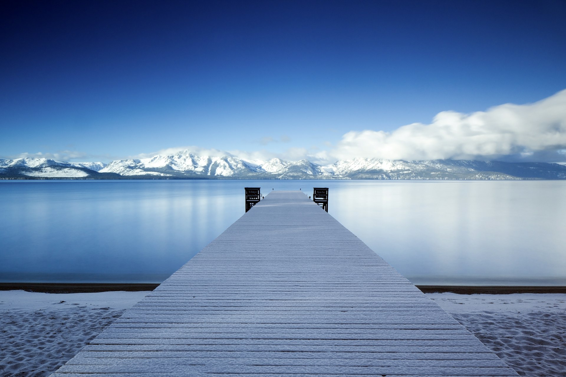A dock protrudes into a flat lake with snowy peaks in the distance