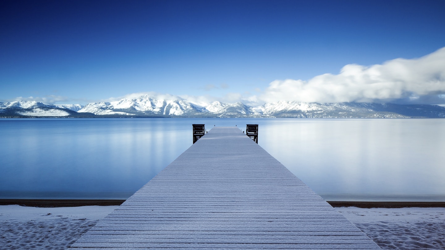 A dock protrudes into a flat lake with snowy peaks in the distance