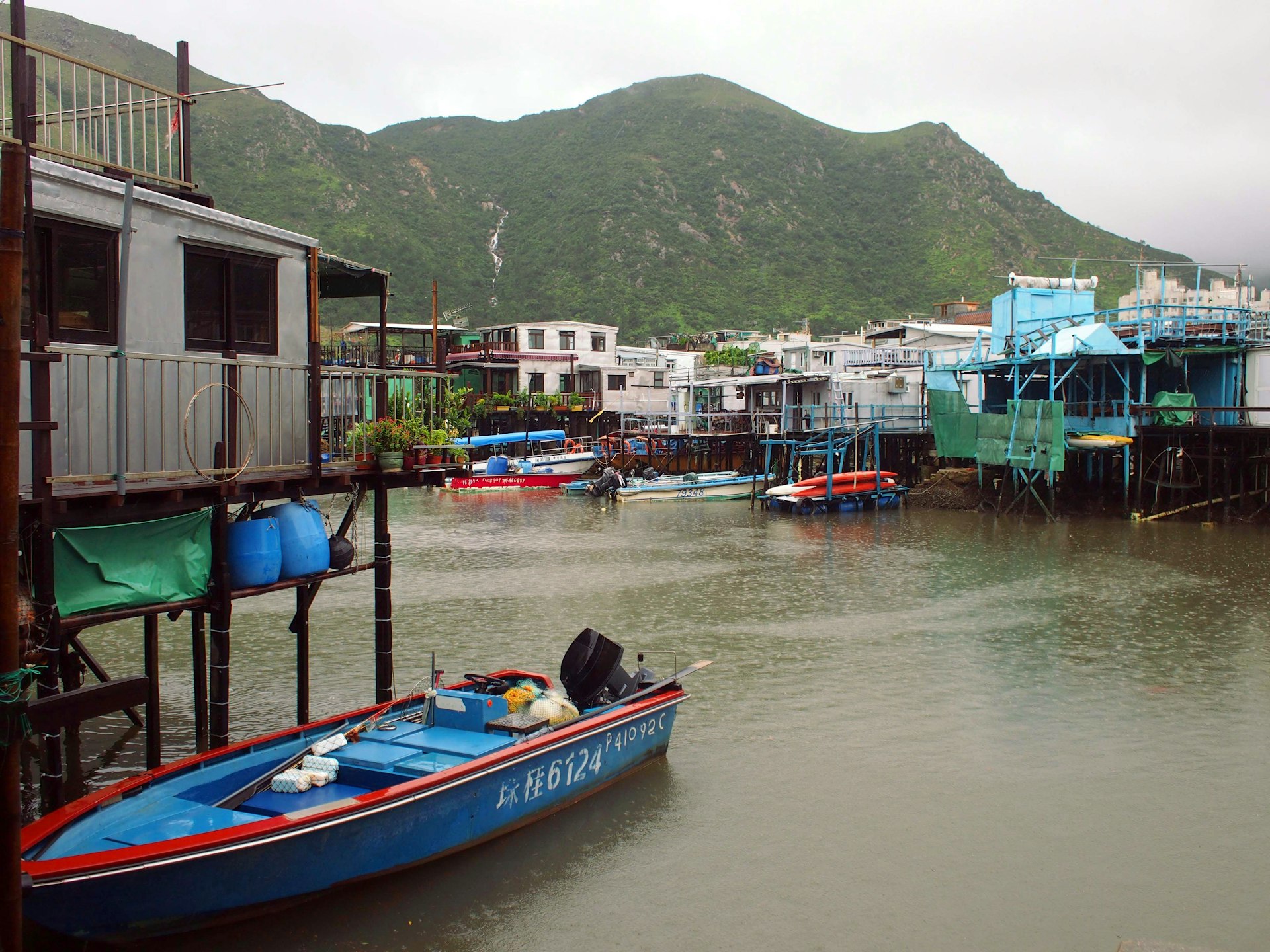 Blue boats sit on a waterway below stilt houses and green mountains