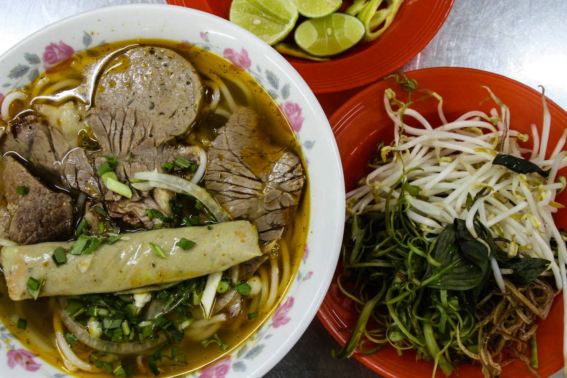 A bowl of spicy beef noodle soup with sides of fresh herbs, vegetables and lime