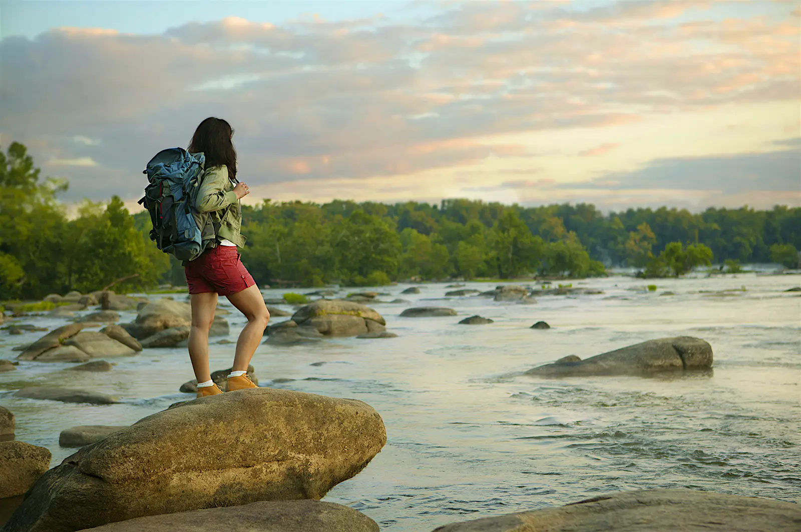 Features - Young woman standing on rock beside river woman with a backpack stands on a rock looking out over the James River at sunset