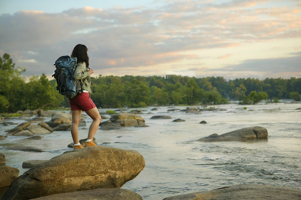 woman with a backpack stands on a rock looking out over the James River at sunset