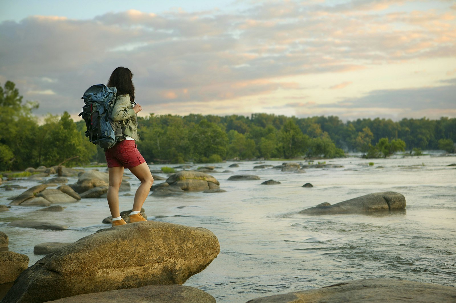 woman with a backpack stands on a rock looking out over the James River at sunset