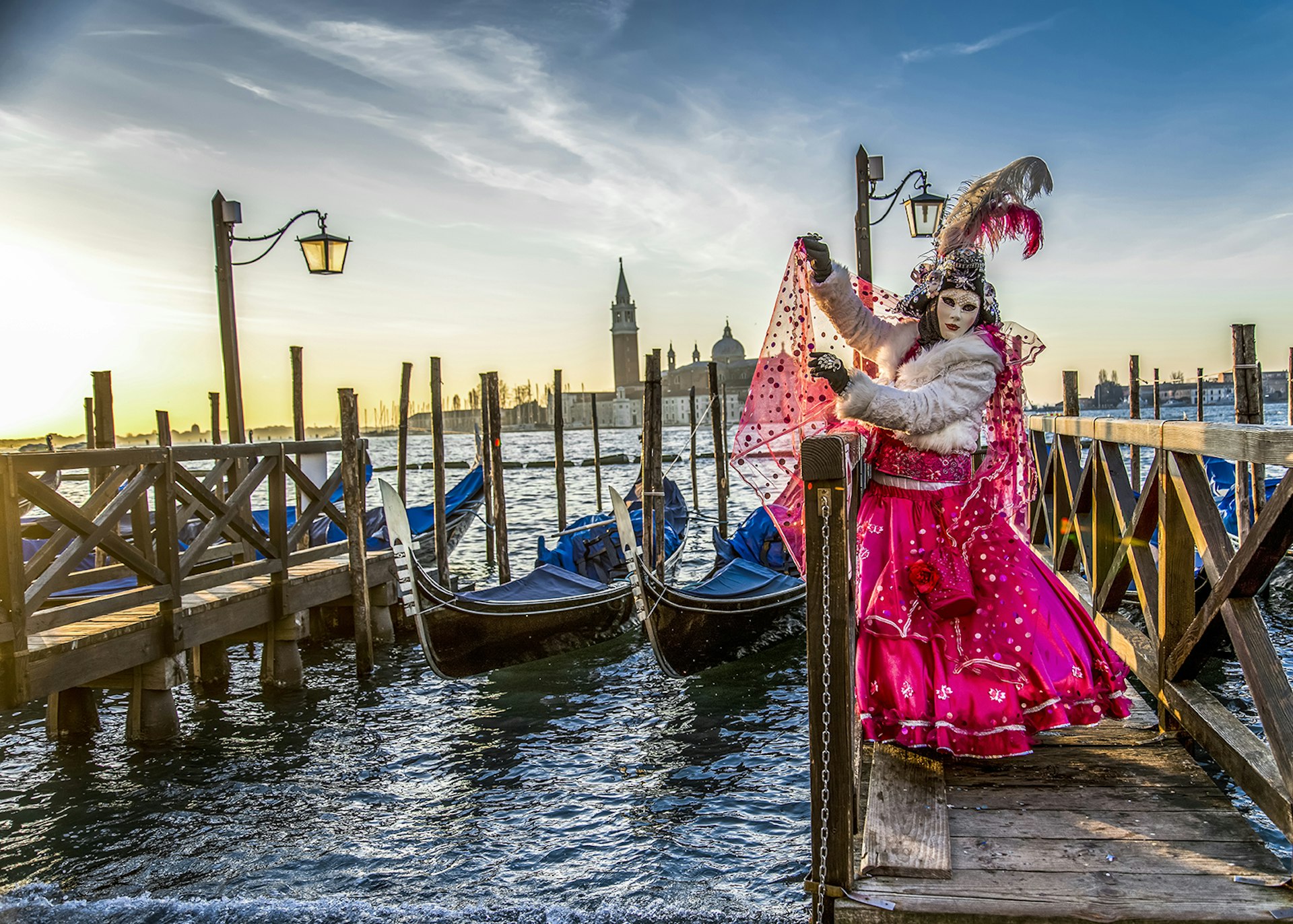 A masked person in a bright pink 18th century Venetian costume throws their wrap into the wind over one of Venice's twinkling canals in front of parked gondolas and a setting sun.