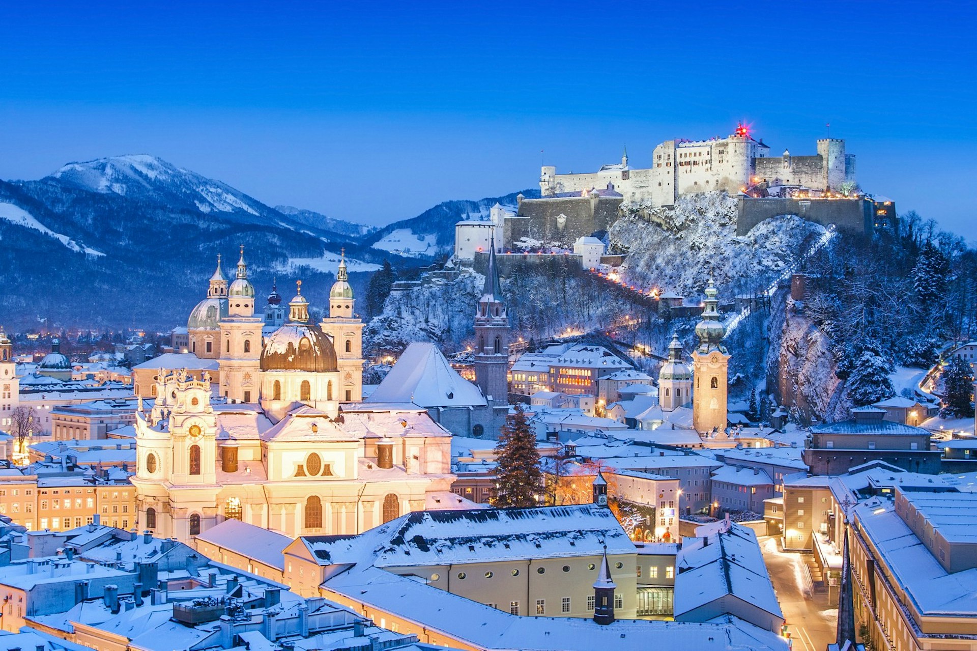 Salzburg Cathedral and Festung Hohensalzburg covered in snow and aglow under evening streetlights