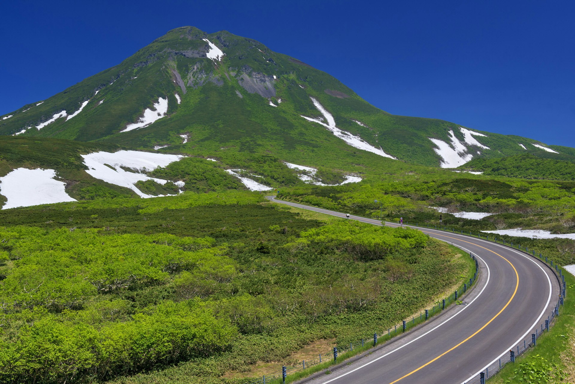 A road leads through a lush green landscape towards mountain Rausu-dake