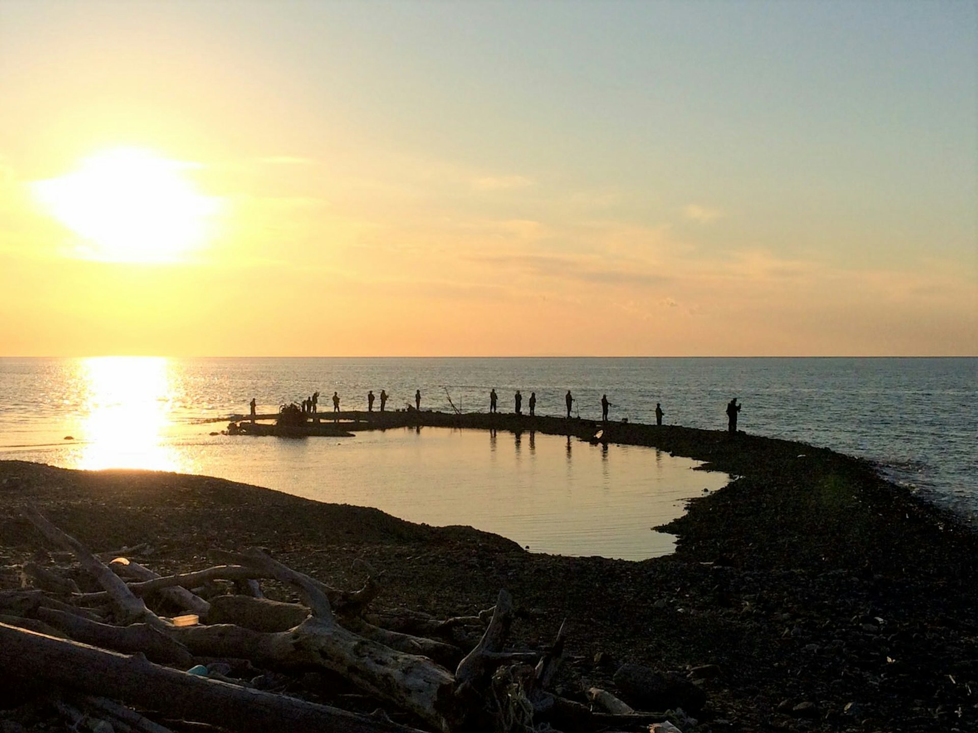 A dozen people stand on a small rocky outcrop fishing as the sun sets over the sea