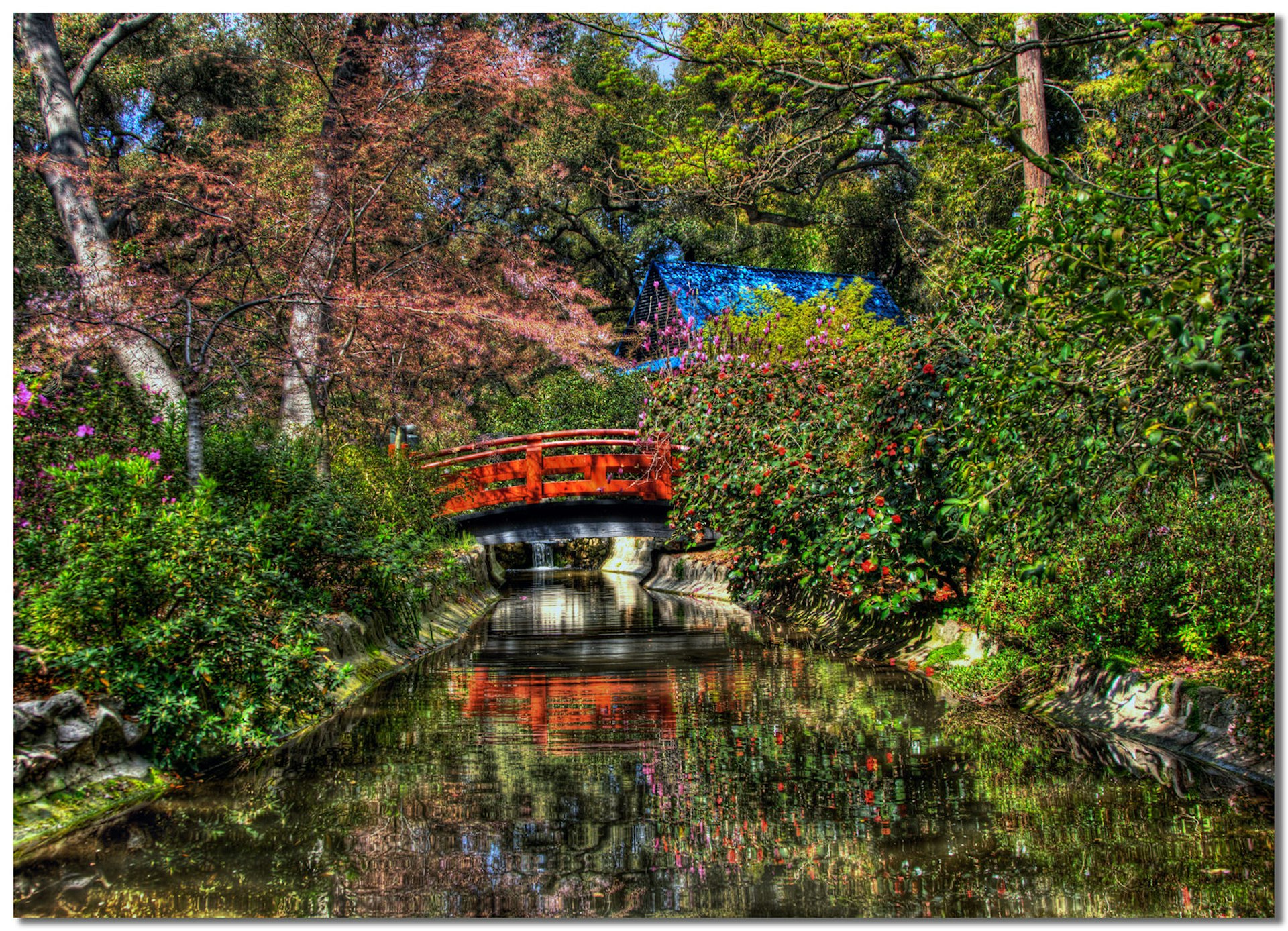 A red japanese-style bridge is reflected in a stream and surrounded by colorful flowers and shrubs.