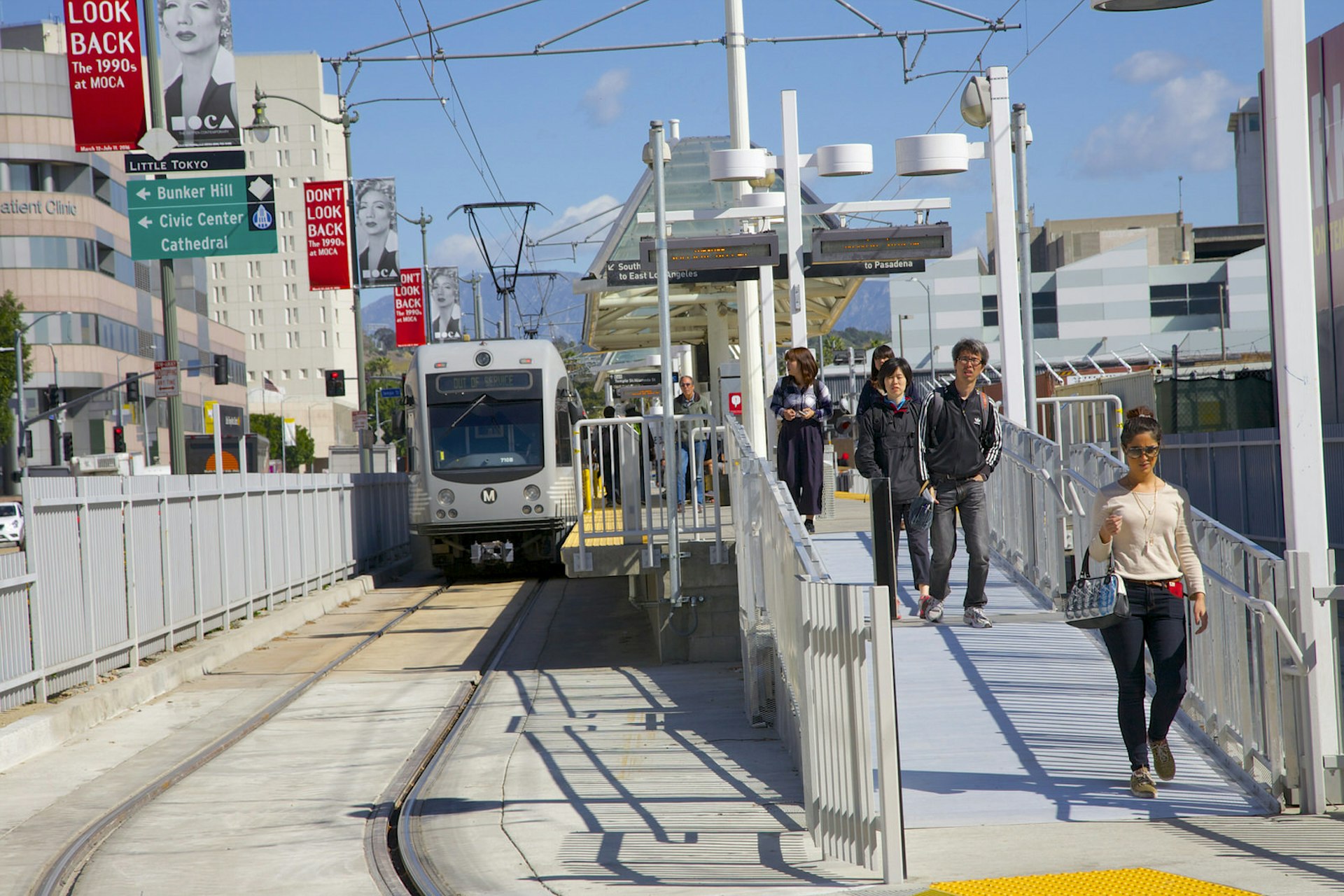 A light rail train sits at the Little Tokyo/Arts District station on the Gold Line. A line of pedestrians walks away from it on the right.