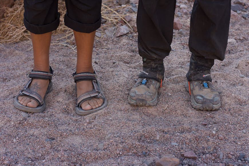 En beduinguide - klädd i ett par välslitna sandaler - står bredvid en vandrare i vadderade vandringskängor © Jen Rose Smith / Lonely Planet