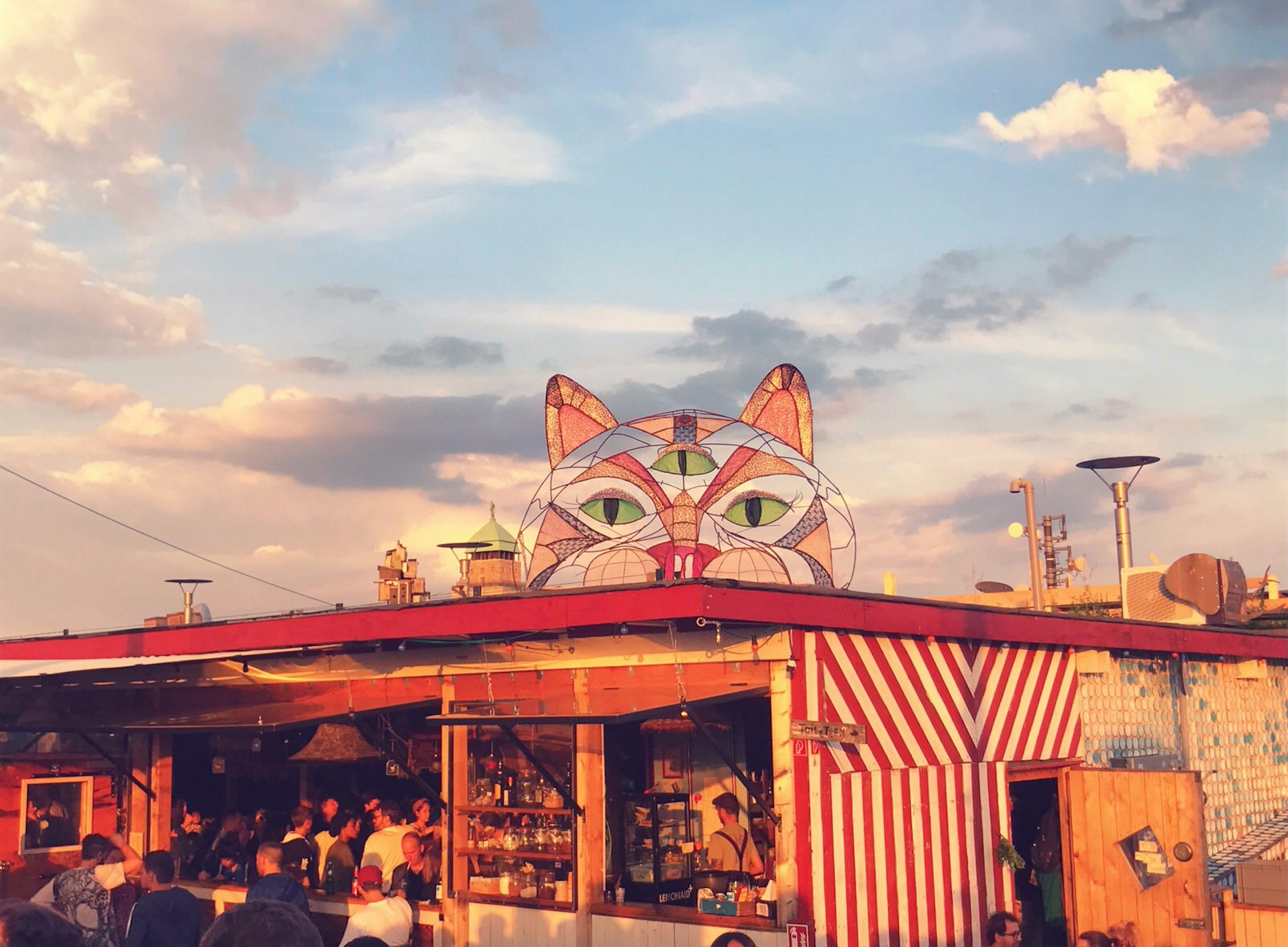 A colourful three eyed cat casts its gaze over the rooftop housing the bustling bar at Klunkerkranich against a cloudy blue sky. © Jennifer Sojka / Lonely Planet