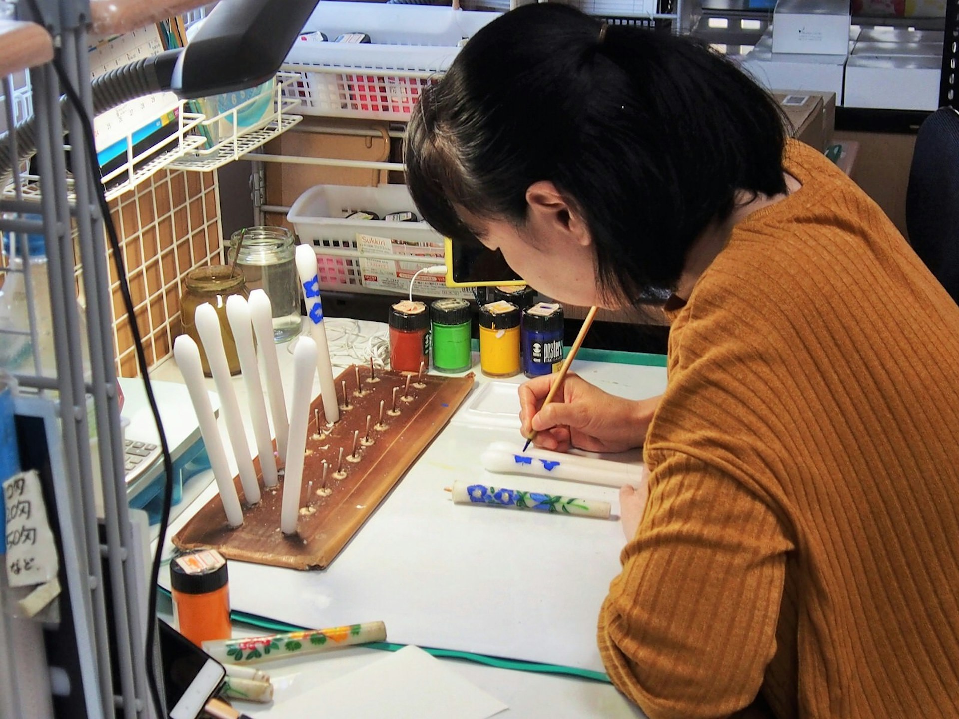 An artist sits at a small workshop desk using a fine brush to paint purple flowers onto a candle © Manami Okazaki / Lonely Planet
