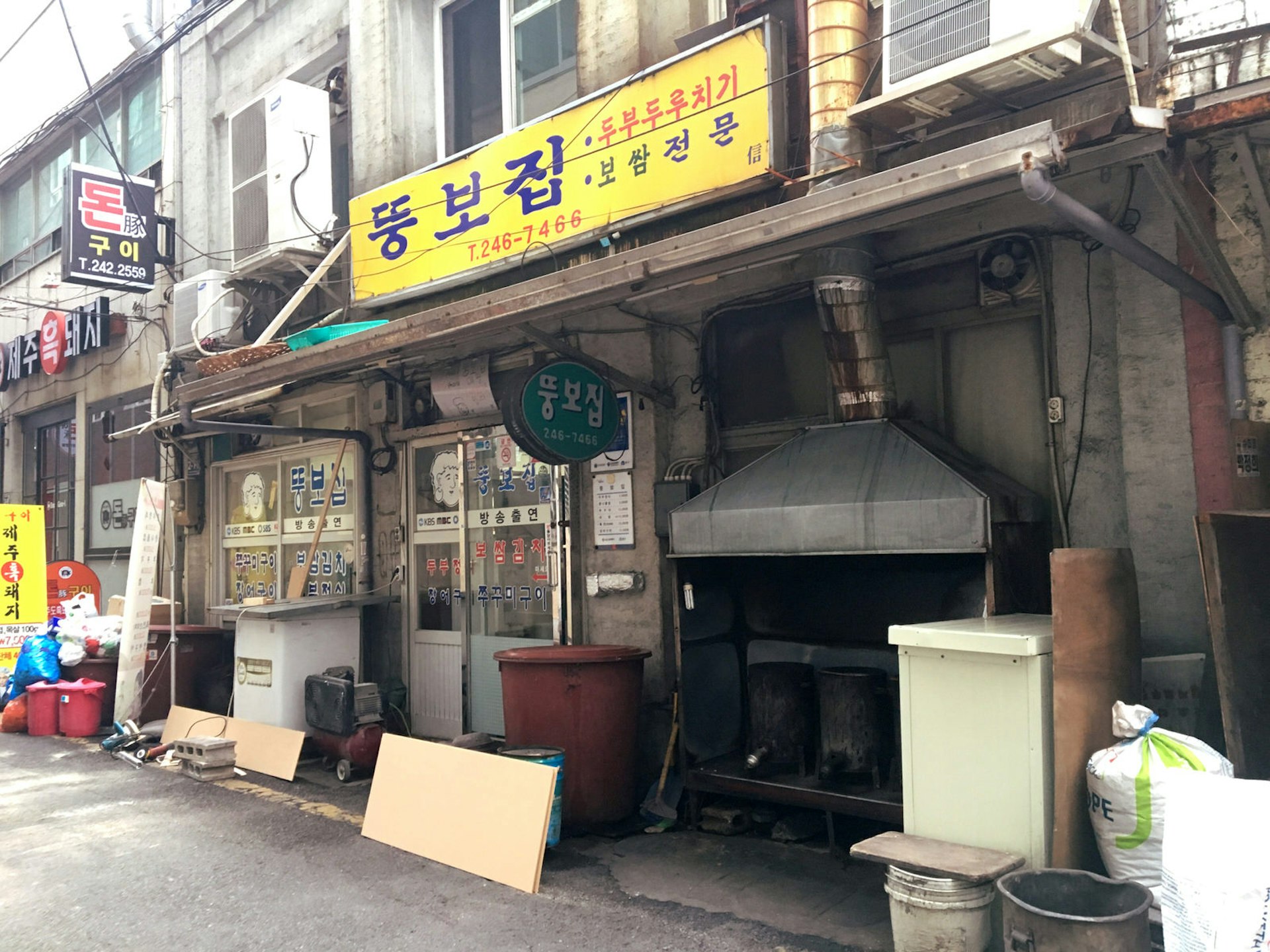 A yellow sign in Korean marks the entrance to Ddoongbo Jip, where a large metal coal-fire grill sits beside the glass doors © Hahna Yoon / Lonely Planet
