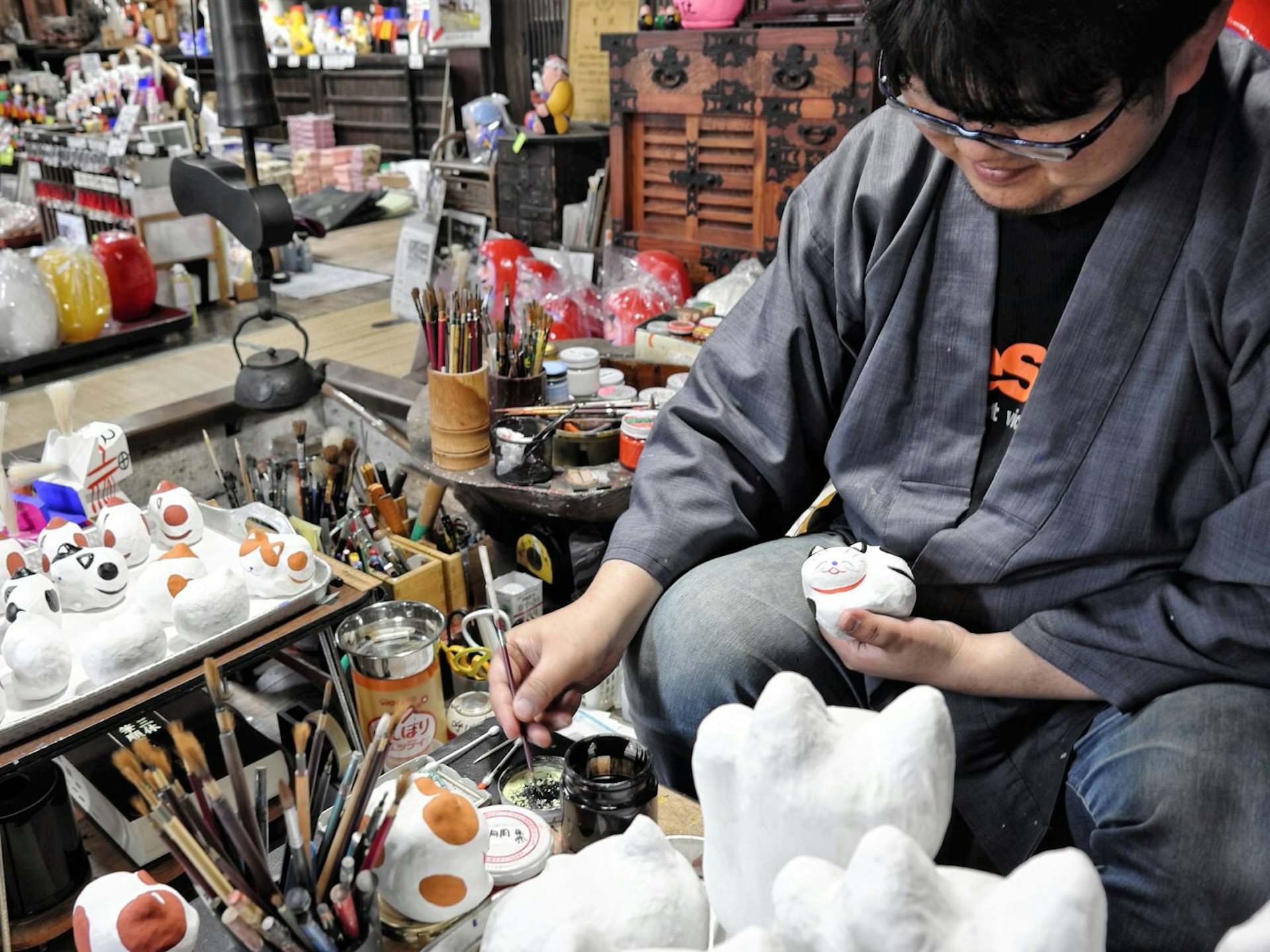 Artisan Daisuke Hashimoto sits in a studio surrounded by pains and various works in progress, painting a small papier mache cat © Manami Okazaki / Lonely Planet
