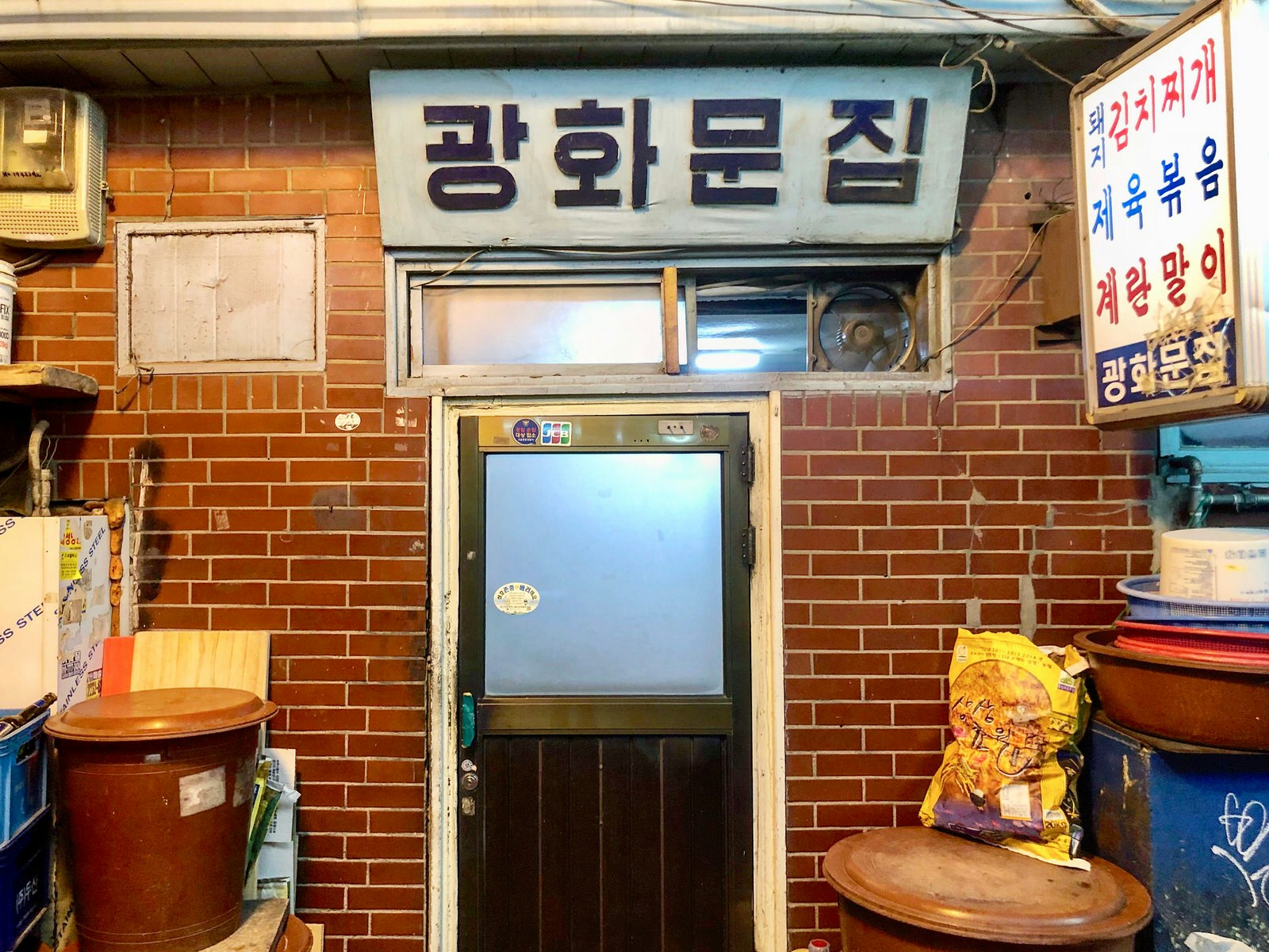 The door of Gwanghwamun Jip in Seoul with a sign in Korean above it and several bins to each side along a brick wall © Hahna Yoon / Lonely Planet