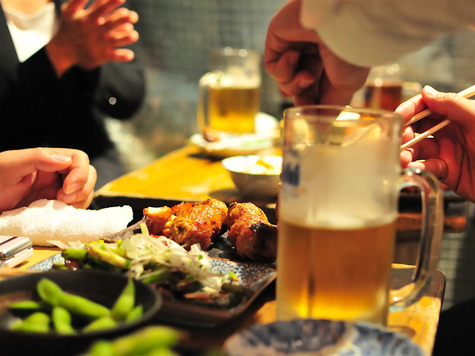 Features - Izakaya Close up of small dishes of grilled meat and vegetables on a table, with half-full glasses of beer and people eating © Photos from / Getty Images