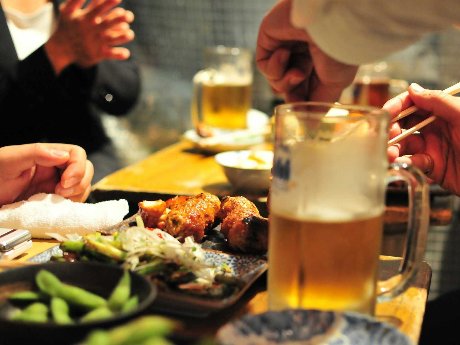Close up of small dishes of grilled meat and vegetables on a table, with half-full glasses of beer and people eating © Photos from / Getty Images