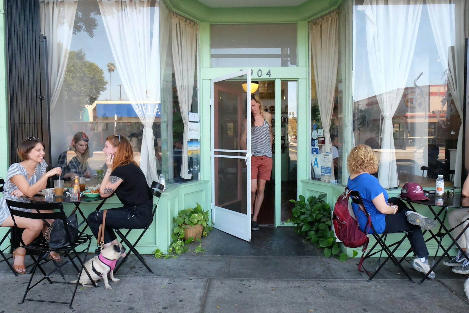 People walk in and out of a cute cafe, with people sitting outside at sidewalk tables.