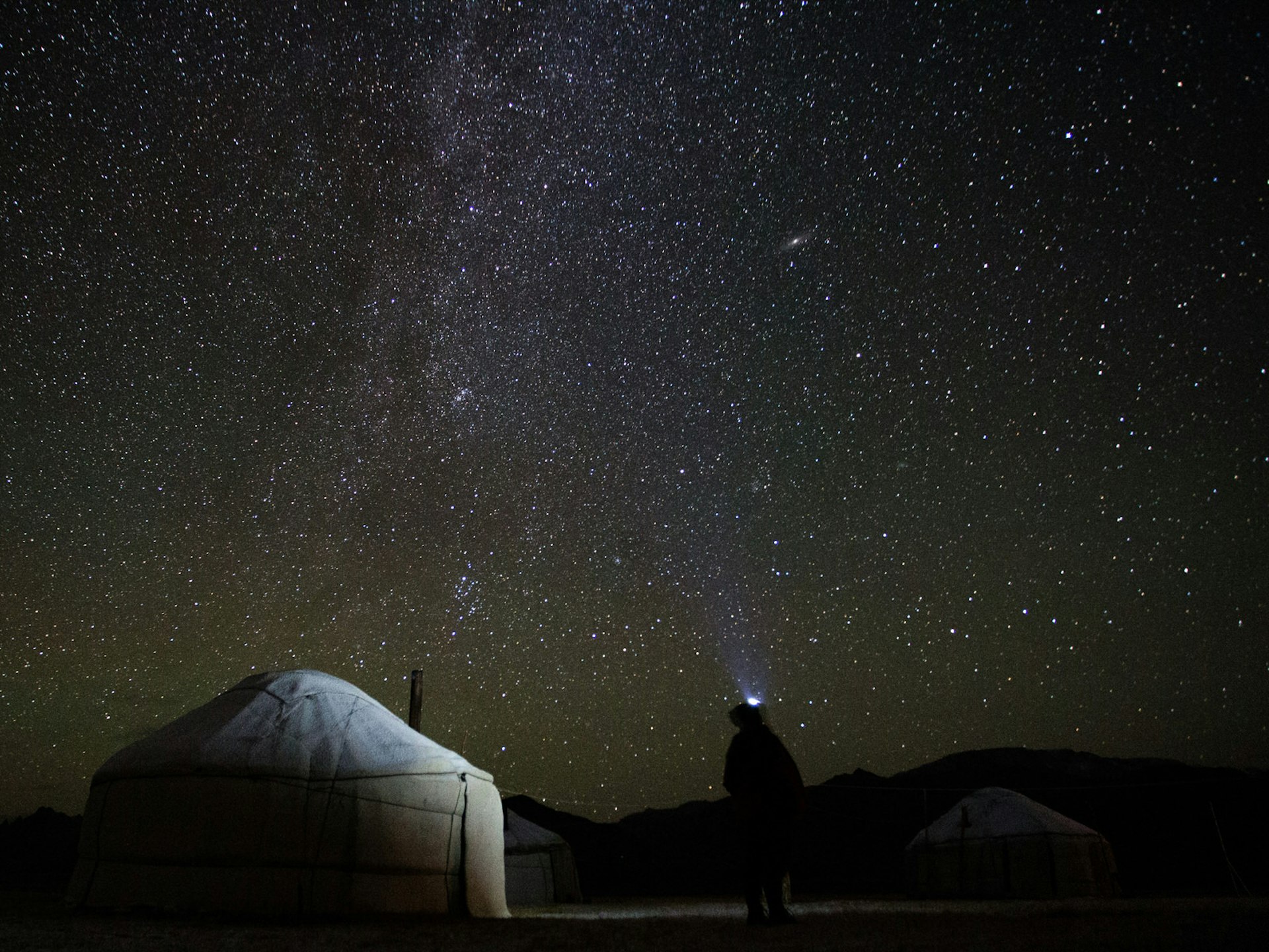 Night sky full of stars. In front, a semi-illuminated yurt tent and a person standing looking upwards with a head torch glowing. © Stephen Lioy / Lonely Planet