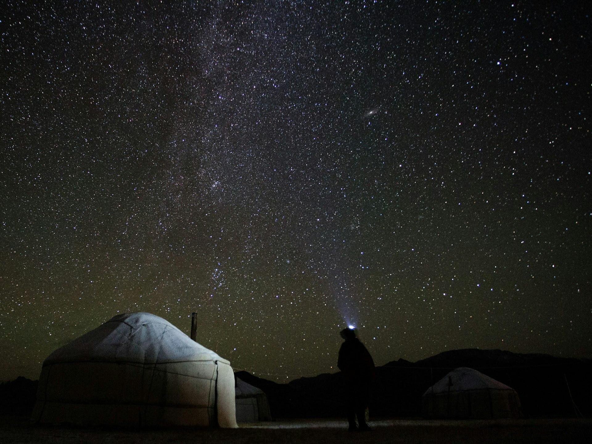 Night sky full of stars. In front, a semi-illuminated yurt tent and a person standing looking upwards with a head torch glowing. © Stephen Lioy / ϲʼʱ