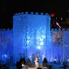 A transparent castle made of giant ice blocks is lit with a blue light from inside, against a pitch black sky.