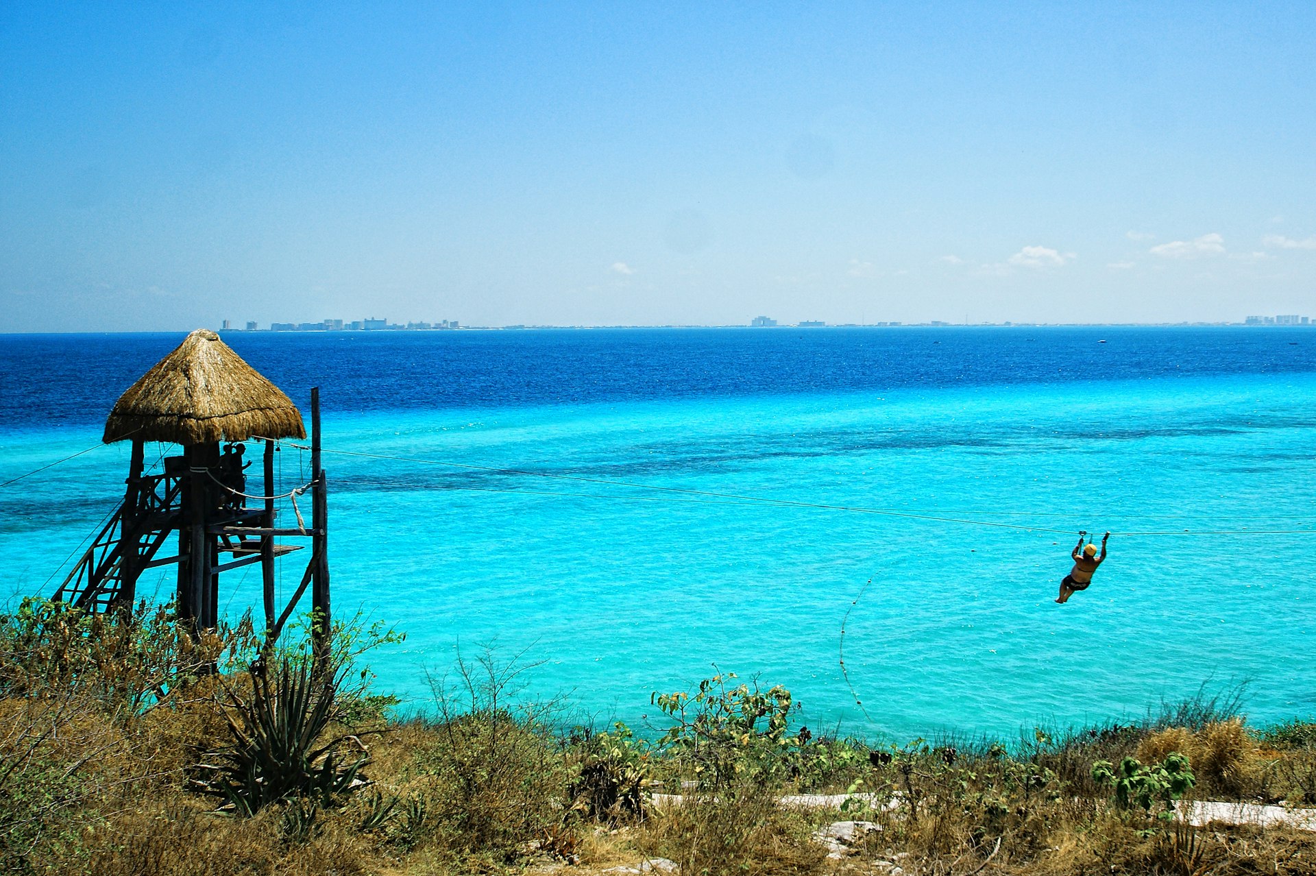 A person ziplines over the Caribbean Sea on Isla Mujeres with Cancún in the background © Infinite Highway / Getty Images