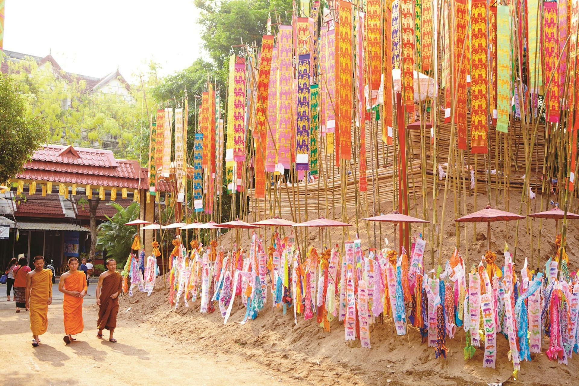 Two monks, wearing bright orange robes, walk past a sand pagoda decorated with colourful prayer flags in Chiang Mai